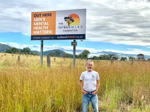 Man standing in a wheat field, in front of a large billboard advertising charity The Outback Mind