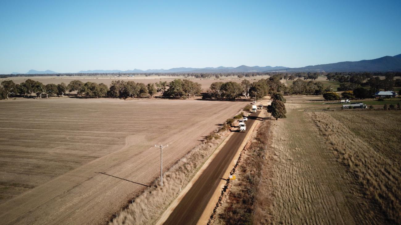 Very long shot of a rural road cutting through a paddock, with construction vehicles driving along it.