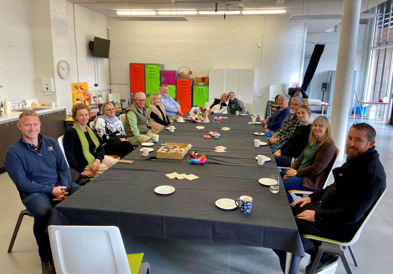 20 people sitting at a long table having morning tea, smiling at camera