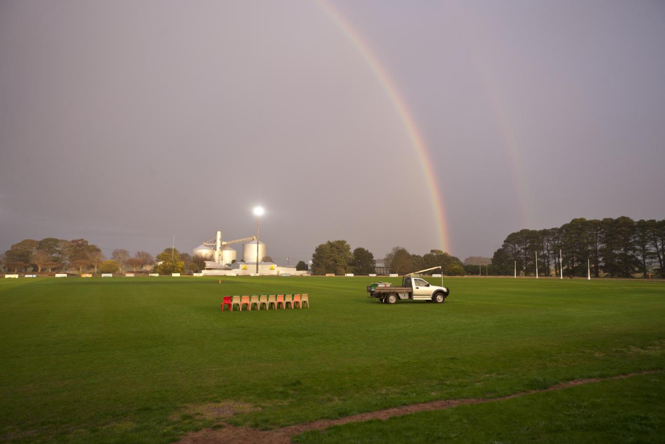 A sports oval in the twilight with a large rainbow curving over the grass and a vehicle closer to the foreground