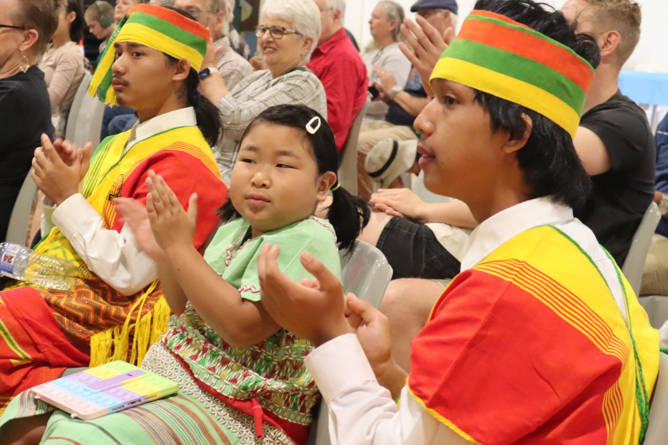 Two young men in traditional dress with young girl, all are clapping while watching a performance