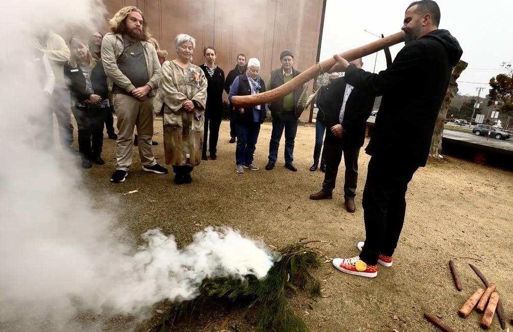 An indigenous man playing didgeridoo at a Smoking Ceremony outside with many people in attendance