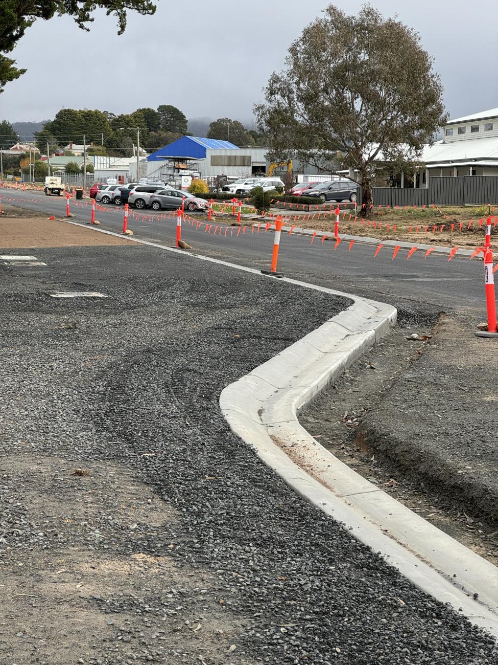 A road in the process of being resealed, with work vehicles and witches' hats on the ground.