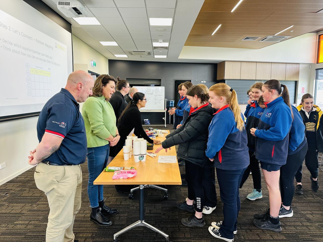 A group of people; a trestle table in the middle of the room with a group of adults on one side and a group of engaged young students on the other