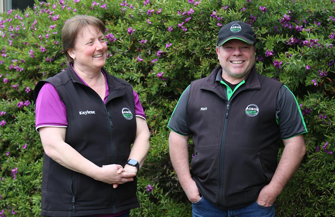 A man and a woman smiling and standing in front of a bush with purple flowers