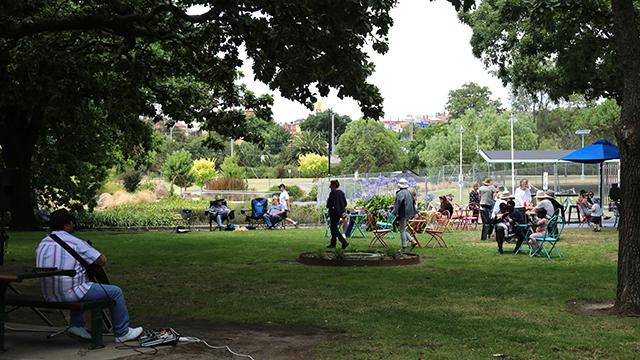 A long shot of a green parkland space with people sitting on picnic chairs and a man playing guitar