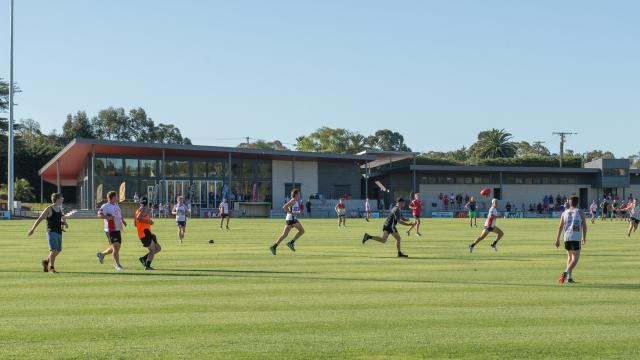 Men playing a game of Australian rules football on a grassy field, with a building and trees in the background.